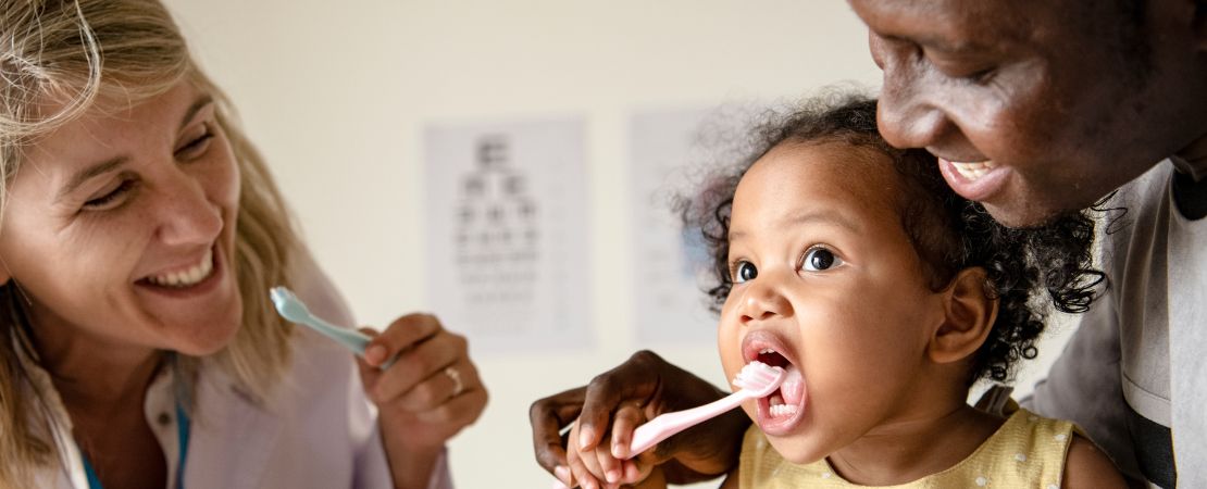 A smiling healthcare professional and a parent help a young child brush their teeth.